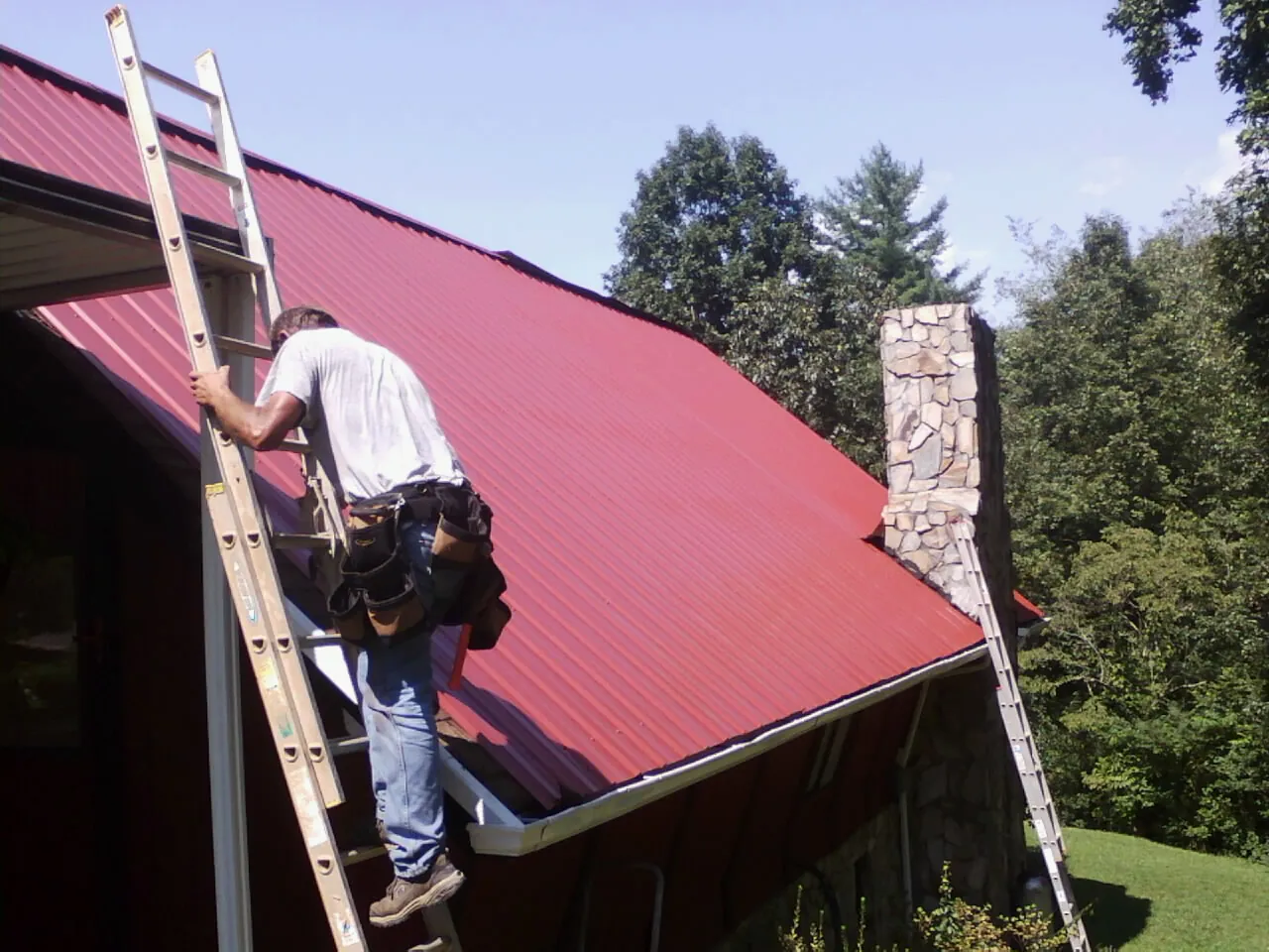 Roofer on a ladder beside a red metal roof and stone chimney — Soffit & Fascia Repair by Roofing Brothers in Westfield
